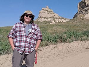 Taylor Green, wearing a plaid shirt and gray pants, stands in a western Nebraska landscape